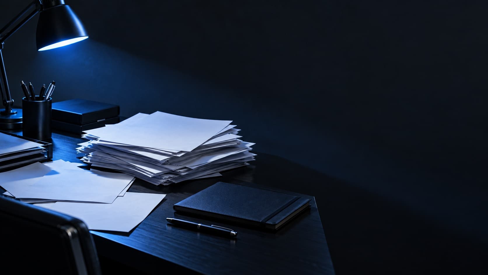 Mesa escura de escritório com papéis impressos empilhados de forma desorganizada, um caderno fechado e uma caneta.